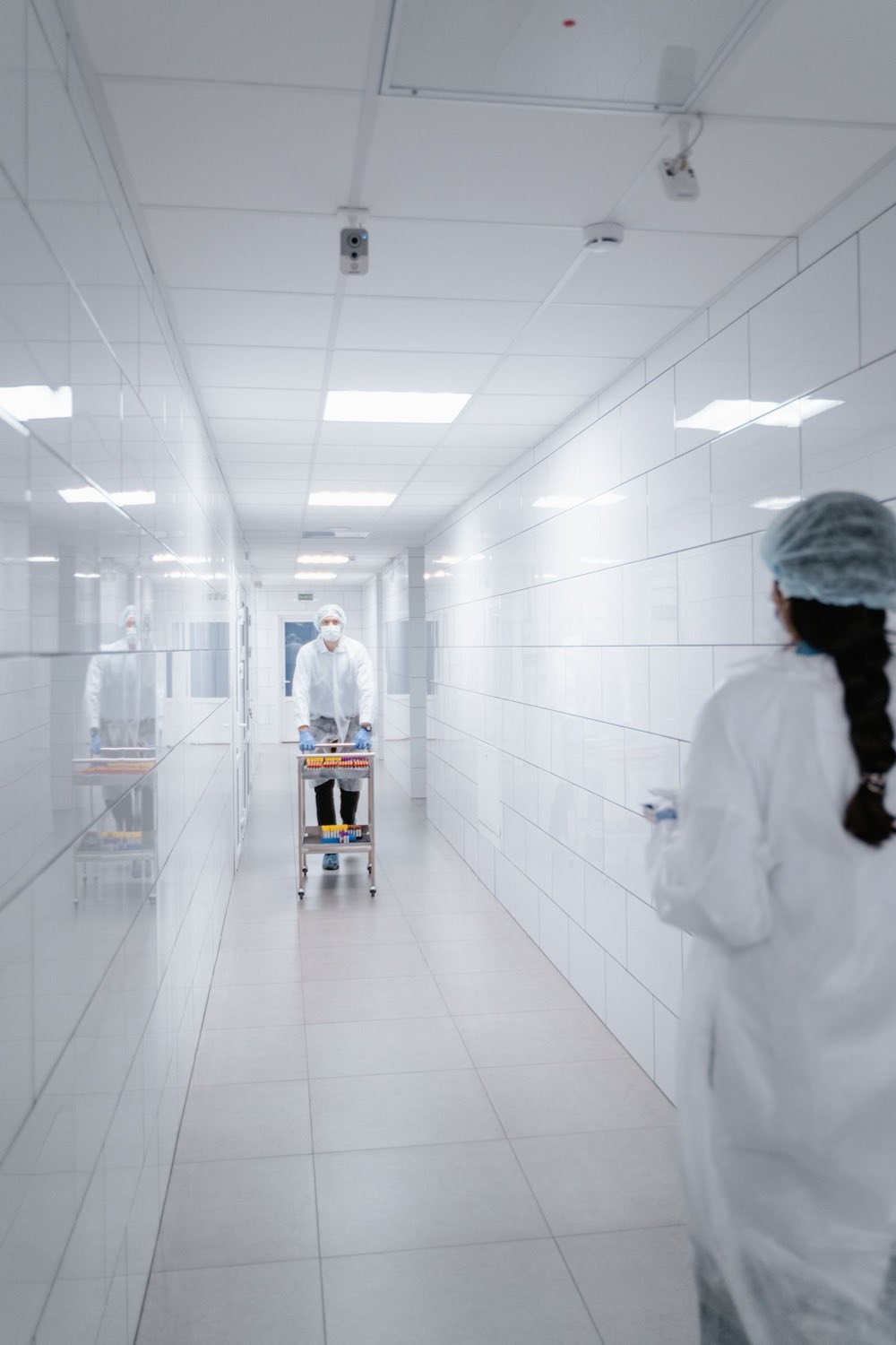 Gowned pharmaceutical operators moving a cart of sample vials through a cleanroom corridor between airlocks