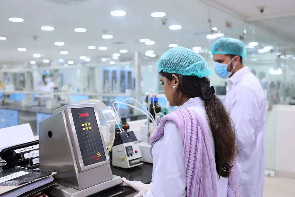 Technicians in hair nets and lab coats working in a pharmaceutical quality control laboratory behind glass partitions